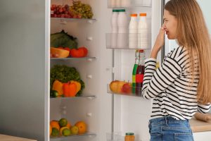 Woman holding her nose in front of a fridge, indicating spoiled food due to fridge not cooling.